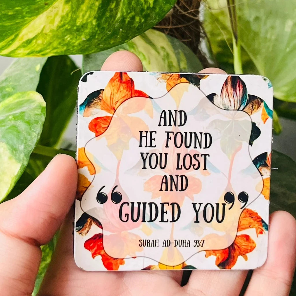 A square metal wall plaque held in a hand, featuring a floral design with orange, yellow, and black accents, and the text 'AND HE FOUND YOU LOST AND GUIDED YOU' SURAH AD-DUHA 3:57'.